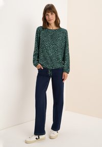 Grünes Langarmoberteil mit einem leichten Muster, kombiniert mit marineblauen Hosen und weißen Sneakern. Das Oberteil hat einen runden Ausschnitt und einen Kordelzug in der Taille.