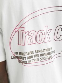 Hvid bomulds-t-shirt med pink grafisk tekst "Track C" og sort tekst, der omhandler temaer som fællesskab og ensomhed. Korte ærmer, afslappet pasform.