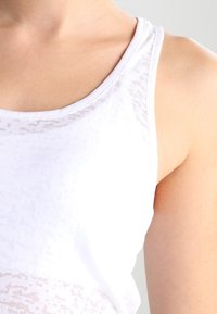 Biały top na ramiączkach o gładkiej fakturze z delikatnymi wzorami, zaokrąglonym dekoltem i szerokimi ramiączkami, zaprojektowany dla wygodnego dopasowania.