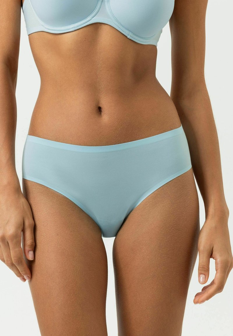 Frau trägt hellblauen nahtlosen BH und passende Unterwäsche, zeigt Bauchpartie und obere Oberschenkel vor einfachem Hintergrund.