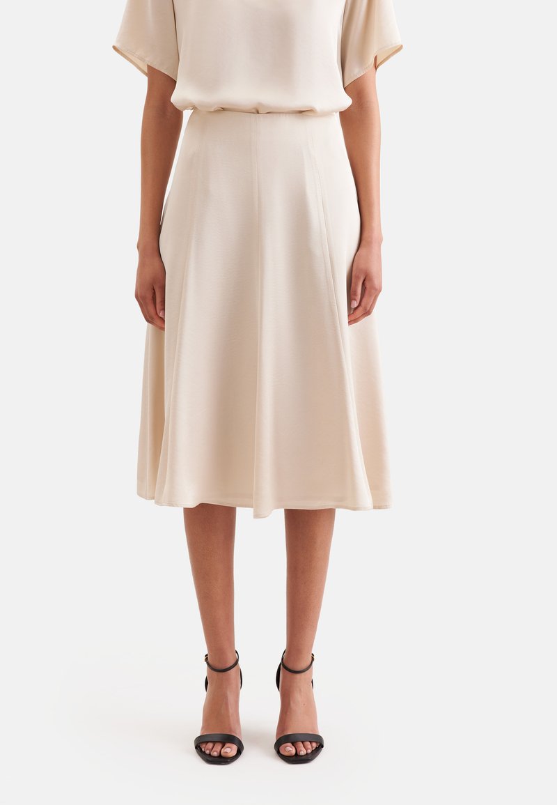 Femme portant une jupe midi beige et un chemisier assorti, associée à des talons noirs ouverts à la cheville, debout devant un fond uni.