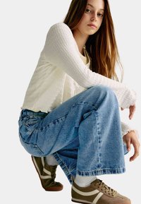 Top de manga larga texturizado blanco, camiseta amarilla clara debajo, jeans de pierna ancha azul deslavado, zapatillas marrones con rayas y cordones crema.