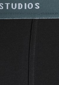Black ribbed fabric with a soft texture features an elastic waistband in green, labeled with “STUDIOS” in white lettering.
