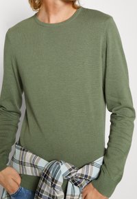 Olijfgroen trui met een ronde hals en lange mouwen, gemaakt van een gestructureerde brei. Een geruite flanellen shirt geknoopt om de taille in blauw en wit.