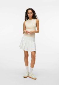 Top senza maniche lavorato a maglia di un leggero beige con motivo testurizzato e piccolo logo nero, abbinato a una gonna plissettata bianca e sneakers beige.