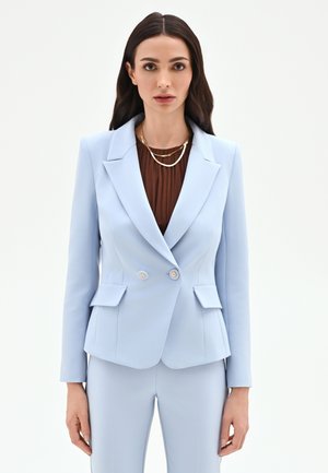 Femme aux longs cheveux foncés portant un costume bleu clair cintré et des colliers en couches dorés sur un haut marron, sur fond blanc.