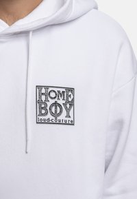 Balta kapuce ar mīkstu tekstūru, kurai ir taisnstūrveida melns logo ar uzrakstu "HOME BOY loud-couture" stilizētām burtiem un faktūrām.