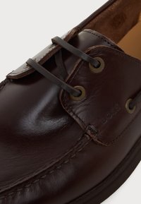 Gros plan sur une chaussure habillée en cuir marron foncé avec des lacets en cuir passés à travers des œillets en laiton et un logo BOSS embossé sur le côté.