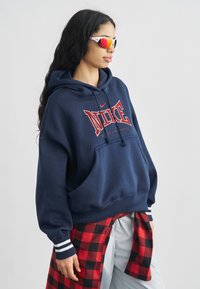 Μπλε ναυτικό φούτερ με κόκκινο λογότυπο "NIKE", κορδόνι και μπροστινή τσέπη. Διαθέτει ριγέ μανσέτα σε μπλε ναυτικό και λευκό. Κόκκινο και μαύρο καρό πουκάμισο δεμένο γύρω από τη μέση.