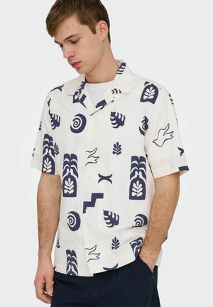 Jeune homme portant une chemise blanche à manches courtes avec des imprimés abstraits et botaniques bleu marine, associée à un pantalon foncé, regardant vers le bas.