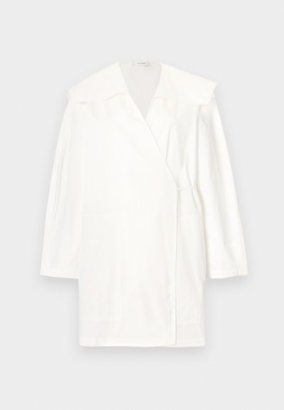 Άσπρο φόρεμα με μακριά μανίκια τύπου wrap, ευρύ γιακά και διακριτική λεπτομέρεια δέσιμου στη μέση, σε ανοιχτό γκρι φόντο.