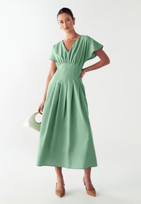 Un vestido fluido de longitud a la rodilla en verde menta, con mangas cortas, falda plisada y cintura ajustada. Se lleva con tacones estampados y un bolso blanco.