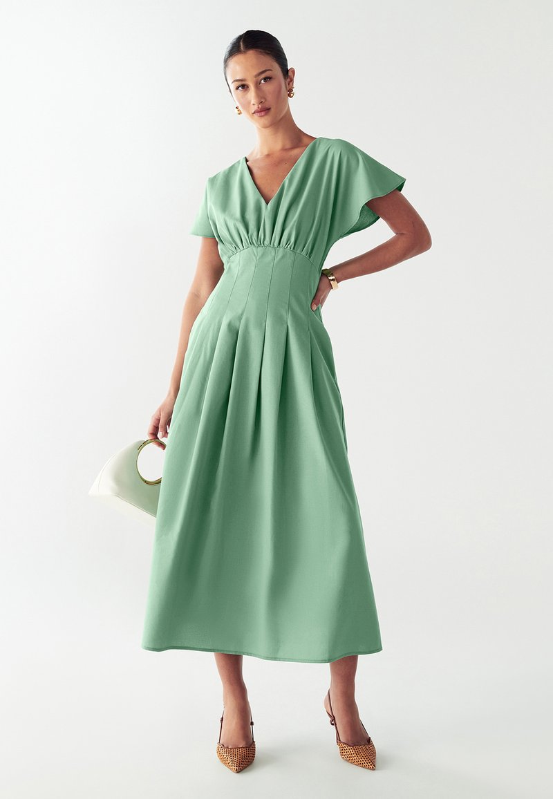 Un vestido fluido de longitud a la rodilla en verde menta, con mangas cortas, falda plisada y cintura ajustada. Se lleva con tacones estampados y un bolso blanco.