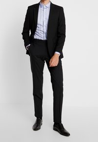 Muž v černém obleku s světle modrou pruhovanou košilí a černými společenskými botami, stojící s jednou rukou v kapse.