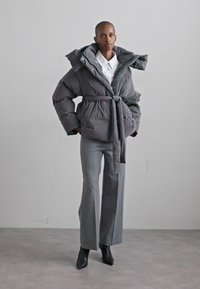 Grey puffer jacket with a belted waist, oversized hood, and buttoned closure, paired with grey wide-leg trousers and black heeled boots.
