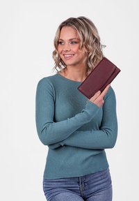 Brown leather wallet held by a person wearing a ribbed teal sweater against a plain white background. Wallet features a smooth texture.