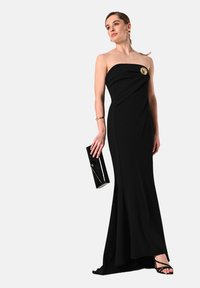 Frau in einem schulterfreien schwarzen Abendkleid mit goldener Brosche, die eine schwarze Clutch hält, goldene Armbänder und Ohrringe trägt und schwarze hochhackige Sandalen mit Riemchen trägt.