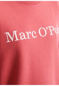 Roosa puuvillane särk; esiküljel on valge trükitud tekst "Marc O'Polo"; sile tekstuur ja standardne ümmargune kaeluskujundus.