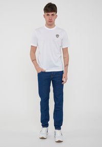 T-shirt bianca in cotone con scollatura rotonda e maniche corte, caratterizzata da un piccolo emblema blu e oro. Abbinata a jeans di denim blu e sneakers bianche.