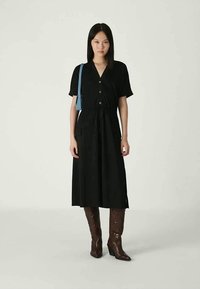 Vestido preto de manga curta com botões à frente, cintura ajustada e saia até ao joelho. Combinado com botas de cowboy castanhas e uma mala azul.