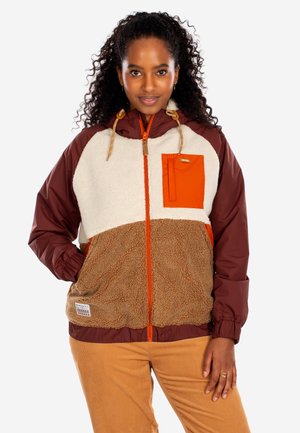 Frau trägt eine Fleecejacke mit Kapuze in den Farben Creme, Braun und Orange, posiert mit Hand in der Tasche und lächelt.