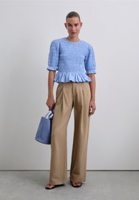 Woman in a textured light blue blouse, beige wide-leg trousers, holding a blue handbag, standing on a gray floor against a white wall.