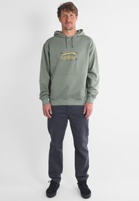 Haut en coton vert avec cordon de serrage, poche avant et un graphique "Oxbow" jaune. Assorti avec un pantalon gris foncé et des baskets noires.