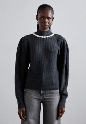Frau mit geflochtenem Haar, die einen dunkelgrauen Rippstrickpullover mit puffigen Ärmeln und einer Perlenkette trägt, kombiniert mit hellgrauen Jeans und steht.