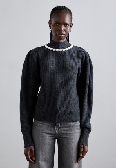 Femme aux cheveux tressés portant un pull à côtes gris foncé avec des manches bouffantes et un collier de perles, associé à un jean gris clair, debout.