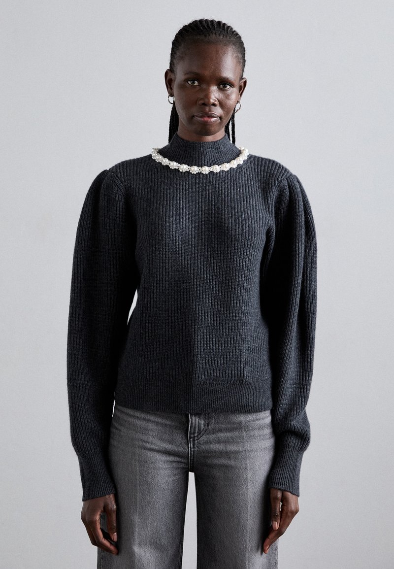 Femme aux cheveux tressés portant un pull à côtes gris foncé avec des manches bouffantes et un collier de perles, associé à un jean gris clair, debout.