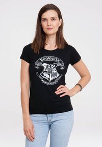 Mustan puuvillapaitojen, joissa on valkoinen Hogwartsin vaakuna -grafiikka. Klassinen malli lyhyillä hihoilla ja pyöreällä kaula-aukolla.
