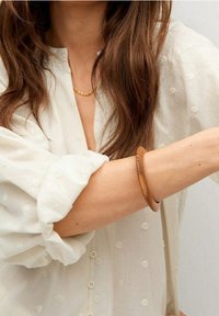 Holzarmreif an einem nackten Arm, mit glatter Oberfläche und warmem Braunton, kombiniert mit einem weißen strukturierten Hemd.