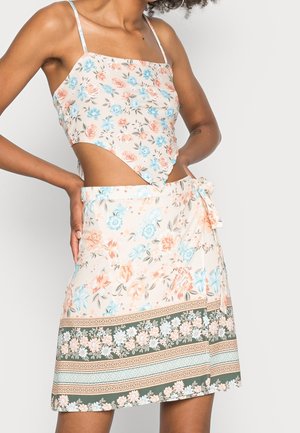 Kobieta ubrana w bezrękawnikowy krótki top w kwiatowy wzór oraz dopasowaną kwiatową spódnicę kopertową w pastelowych odcieniach niebieskiego, brzoskwiniowego i zielonego na jasnym tle.