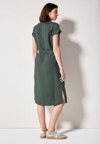 Abito camicia in lino verde con maniche corte arrotolate, spacco laterale e cintura annodata in vita, abbinato a scarpe da ginnastica chiare.