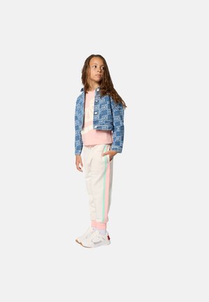 Kind met lang haar, gekleed in een blauwe spijkerjas met patroon, een roze T-shirt, crèmekleurige joggingbroek met pastelkleurige strepen en witte sneakers, staand tegen een effen achtergrond.