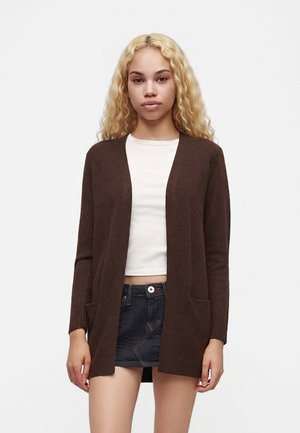 Ung kvinde med langt krøllet blondt hår iført en brun åben cardigan, hvid cropped top og mørk denim mini nederdel.