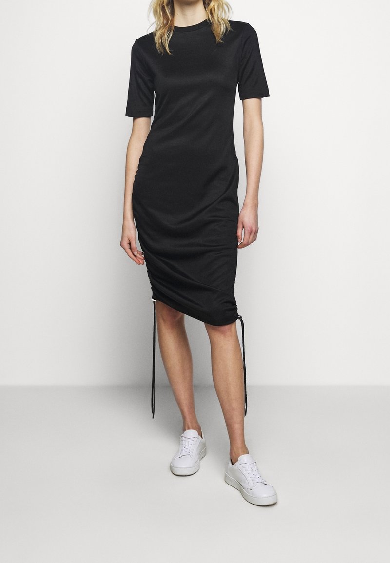 Femme portant une robe midi noire froncée à manches courtes et des baskets blanches, debout devant un fond clair uni.