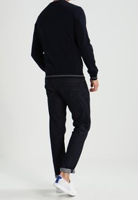 Osoba v tmavém svetru, s vyhrnutými tmavými džíny a bílými teniskami s modrými detaily, stojící na světlém pozadí, čelem k nám.