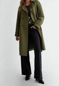 Oliivinvihreä kaksirivinen trenssitakki kauluksella, yhdistettynä mustiin leveneväisiin farkkuihin ja mustiin terävälapaiseen nilkkureihin.