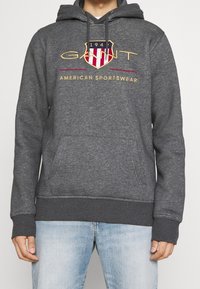 Felpa grigia con tasca frontale, cappuccio con cordoncino e logo scudetto ricamato in rosso, blu e oro, con testo "GANT AMERICAN SPORTSWEAR".