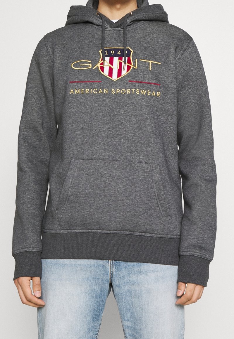 Felpa grigia con tasca frontale, cappuccio con cordoncino e logo scudetto ricamato in rosso, blu e oro, con testo "GANT AMERICAN SPORTSWEAR".