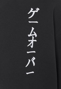 Černá mikina s vertikálním bílým japonským textem. Látka je měkká a texturovaná, má kulatý výstřih a dlouhé rukávy.