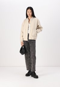 Veste bomber en fausse fourrure crème avec fermeture à pression, deux poches avant, associée à un jean noir à motif étoilé et des bottes chunky noires.