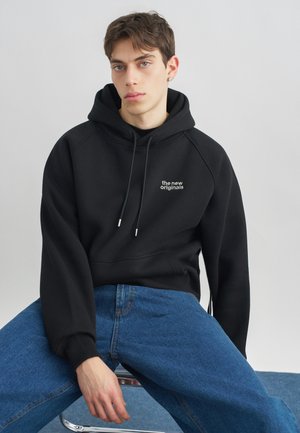 Giovane seduto, indossa una felpa nera con cappuccio con la scritta "the new originals" e jeans blu, su uno sfondo grigio chiaro uniforme.