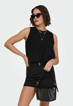 Femme en haut sans manches noir et jupe, portant des lunettes de soleil œil de chat, des colliers argentés, tenant un sac à main noir clouté.