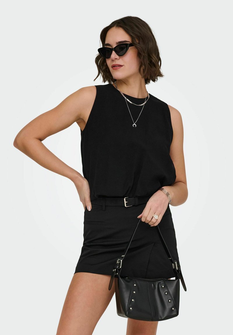 Femme en haut sans manches noir et jupe, portant des lunettes de soleil œil de chat, des colliers argentés, tenant un sac à main noir clouté.