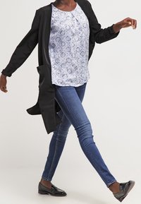 Černý dlouhý cardigan přes bílý top s modrým květinovým vzorem, doplněný úzkými modrými džíny a černými mokasínami. Hladký materiál a přiléhavý střih.