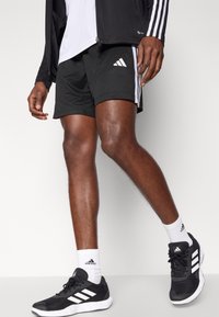 Pantalones cortos deportivos negros con rayas blancas, combinados con zapatillas negras y blancas. Presentan un tejido texturizado y un logo reflectante.