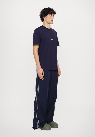 T-shirt blu navy a maniche corte con un piccolo logo frontale, abbinato a pantaloni larghi blu navy con strisce bianche sui lati e calzature verdi.