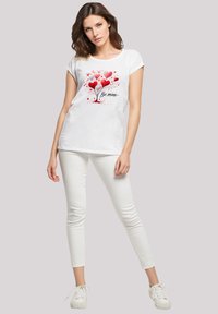 White t-shirt with short sleeves featuring a graphic of red and pink hearts and the text "Be mine" in black. Worn with white skinny jeans.